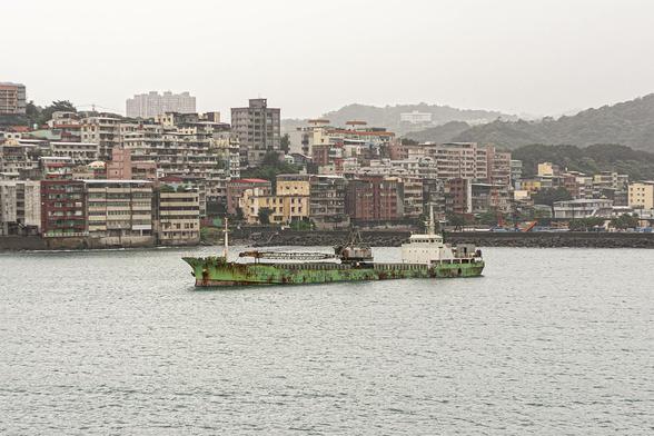It's a murky scene on an overcast day with dull light. We're on the water in a bay approaching a city that lines it, consisting of tightly-packed apartment blocks of various designs, and all with the look of having been there for a few decades, exposed to the elements. A few tree-filled hills rise in the background but they're partly lost to a hazy mist. You can feel a light drizzle in the air even if you can't see it. The water of the bay is flat and grey too, reflecting nothing. Between our position and the shoreline of buildings there is a ship in the water. It's a small cargo carrier and looks to be abandoned, sitting low in the water. Its hull was a bright green once but it's covered in dark, rusty patches. A crane lies along its length but it too looks too rusted to trust to ever lift anything on or off the ship's deck.