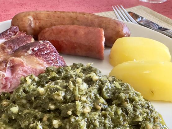 Ein Teller mit geschnittener Wurst, einem Stück Fleisch, gekochten Kartoffeln und einer Portion grünem Gericht, wahrscheinlich eine Gemüse- oder Gemüsezubereitung. Der Hintergrund ist eine helle Tischdekoration.

A plate featuring sliced sausage, a piece of meat, boiled potatoes, and a serving of green dish, likely a vegetable or greens preparation. The background is a light-colored table setting.