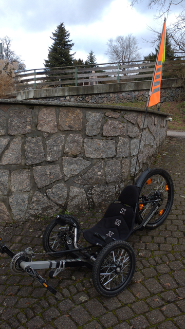 A black KMX X-Class recumbent trike in a drive in front of a natural stone wall. It has a large hi-viz orange security flag sticking out of the backrest. Portrait orientation to get the security flag on the picture, too.