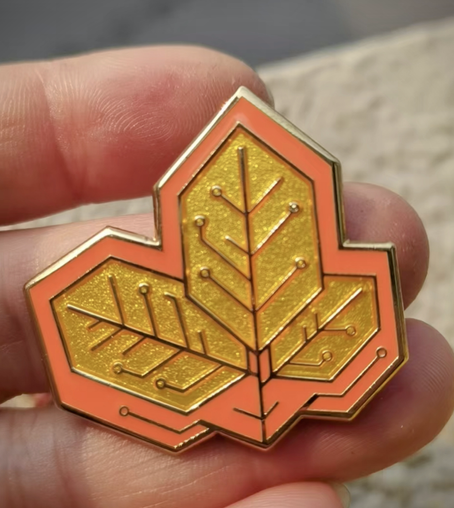 Photo of a hand holding a hybrid enamel pin sample of leaves designed to look like a printed circuit board. This was a colorway test for the Fire Unit Line, and the enamels are a beautiful saturated orange and clear gold.