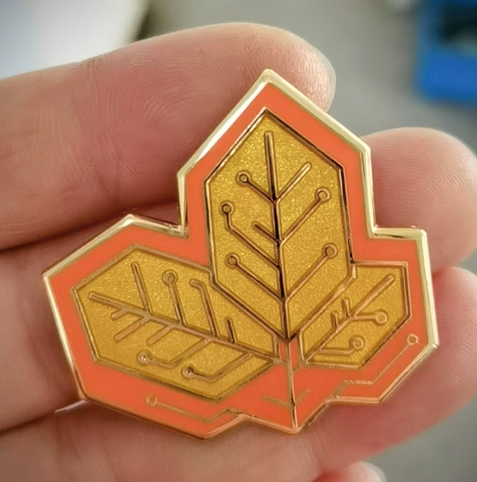 Photo of a hand holding a hybrid enamel pin sample of leaves designed to look like a printed circuit board. This was a colorway test for the Fire Unit Line, and the enamels are a beautiful saturated orange and clear gold.