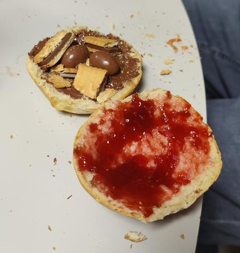 Two halves of a bread roll, one is spread with butter and raspberry jam, the other one with butter, Nutella, PickUp cookies and Kinder Schoko-Bons.