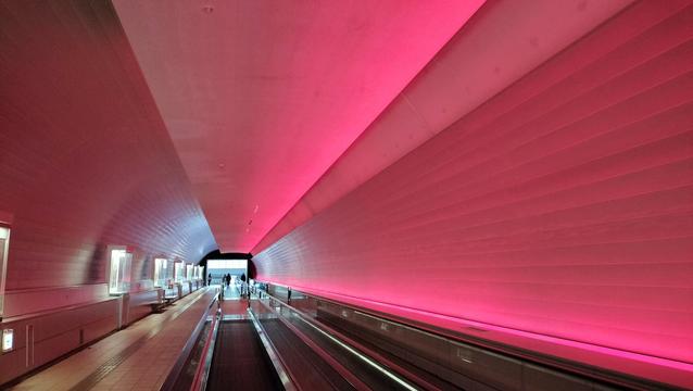 A long pedestrian tunnel. There is a standard walkway as well as moving walkways. The tunnel is vividly lit in red.