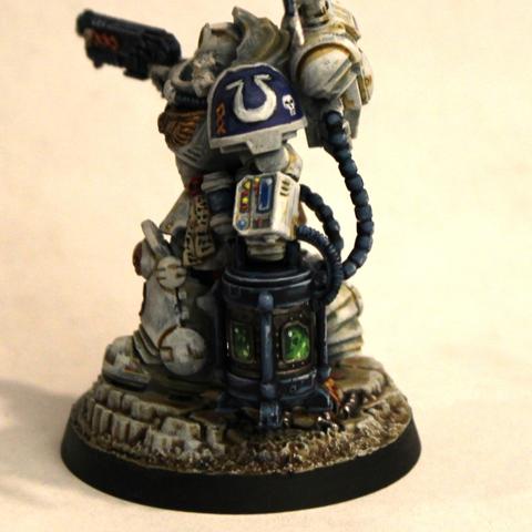Space Marine in white armor with gold wings on the chest. He holds a pistol in one hand and a blue-gray crate with a green glow coming from the windows into it. His armor has lights and vivisection tools on robotic arms and a tool-filled tabard down between his legs. He stands on grimy gray stones and dirt with weird growths and green ooze on it.