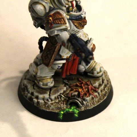 Space Marine in white armor with gold wings on the chest. He holds a pistol in one hand and a blue-gray crate with a green glow coming from the windows into it. His armor has lights and vivisection tools on robotic arms and a tool-filled tabard down between his legs. He stands on grimy gray stones and dirt with weird growths and green ooze on it.