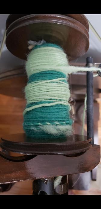 A close-up of the spinning wheel bobbin with dark teal and mint/lime green yarn on it.