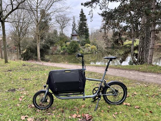 Vélo cargo Muli avec caisse fermée, vue de profil, coté gauche. Il est gris, avec une transmission à courroie sur un moyeu Alfine, une dynamo-moyeu et des éclairage. En arrière plan, le kiosque de l'empereur Napoléon III, au bois de Boulogne.