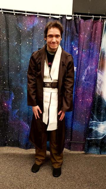 Smiling young man, dressed as a Jedi Knight, stands in front of a curtain backdrop with a starry night sky pattern.