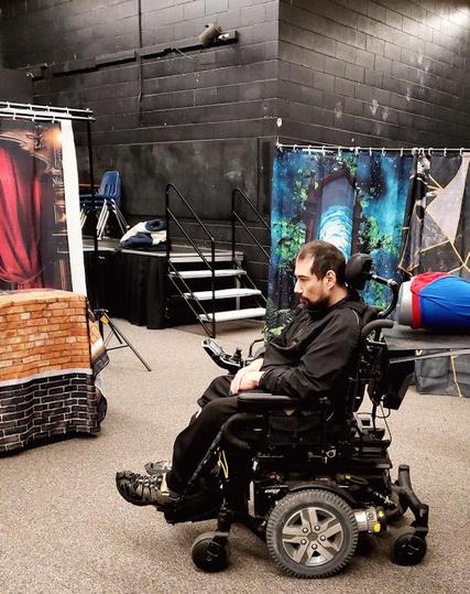 Full body, side view of a young man in a wheelchair - he is in pensive thought contemplating his character for rehearsal. The background shows pieces of set, and a small raised stage area.