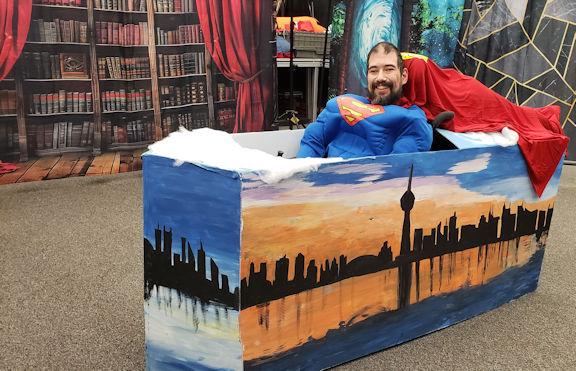 A wheelchair actor is dressed in a superman muscle shirt and cape, the wheelchair is surrounded in a frame depicting the Toronto skyline.
