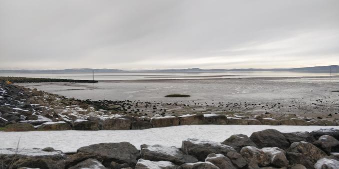 The sea front at Morecambe looking over the bay to the snow-covered hills of the Lake District.

The landscape is almost monochrome.

In the far distance the snowy hills merge into a cloudy sky. The tide is out, the sea is a distant ribbon of light greys.  From the left hand margin a stone jetty sticks out into the mud. In front of that is a groyne made up of dark, snow-spattered boulders. There is a similar line of boulders in the foreground.

It looks as cold as it was.