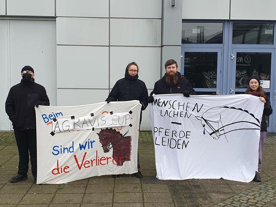 vier leute halten ein banner auf dem steht beim agravis cup sind wir die verlierer mit einem braunen pferd und ein weiteres banner mit der aufschrift menschen lachen pferde leichen, das pferd auf dem banner hat eine träne