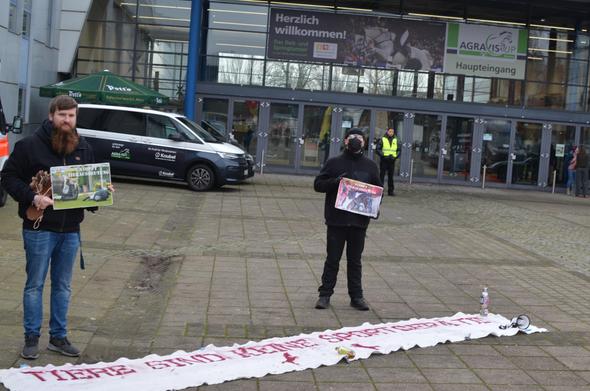 zwei aktivis stehen vor dem eingang der halle münsterlang vor einem banner auf dem steht pferde sind keine sportgeräte, sie halten schilder mit bildern von reitunfällen
