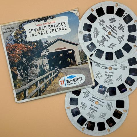 An image featuring a View-Master packaging titled "New England Covered Bridges and Fall Foliage" alongside three View-Master reels. The package shows a covered bridge surrounded by autumn foliage.