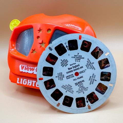 Image features a colorful View-Master toy with a turquoise reel. The reel displays various scenic images labeled "New England" and "Covered Bridges and Fall Foliage." The background is a soft, neutral color.