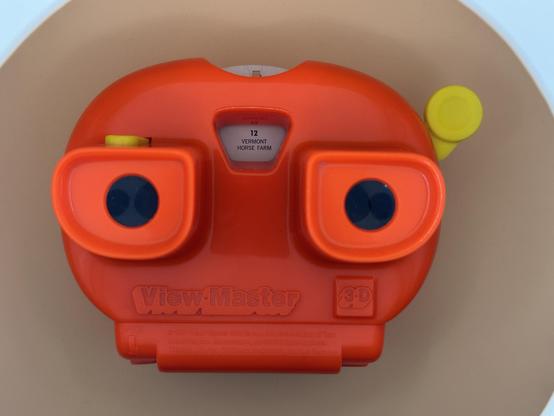An orange View-Master toy with two circular viewing lenses and a label indicating "Vermont Horse Farm" is visible. The device is designed for viewing 3D images.