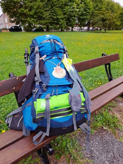 A large blue backpack lying on a bench in a park. A scallop shell is hanging from it by a thread and socks and underware are attached to the bag with clothes pegs.