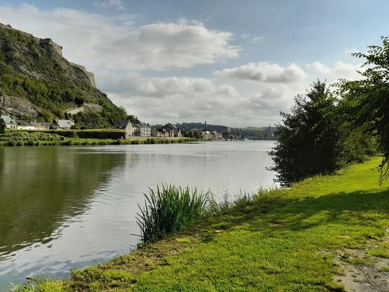 A view of a river with, on the other side to the left a village in front of a hill. On top of the hill part of a fort is visible. To the right, on this side of the river, are grass and some bushes.