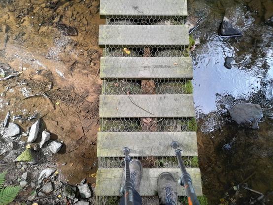 A picture taken while crossing a river, looking down. My feet and hiking poles are just visible and it is clear that the bridge is only wide enough for one person.  There is no handrail. Below the ridge is a very shallow river with some rocks in it.