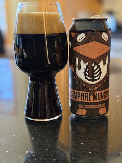A stout glass full of dark beer with a thick tanned. To the right is a pint can with a huge S’more, a light brown graham cracker on top, dripping dark brown chocolate atop a white marshmallow.