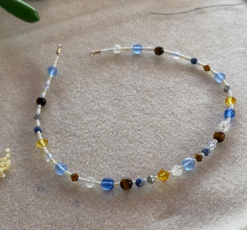 A beaded choker on a cream colored bead mat

The choker is white, silver yellow, gold, blue, and brown. You can see a few inch piece of beading wire, the end of a green tool handle, and a few butter yellow seed beads with a pearl finish on the bead mat.