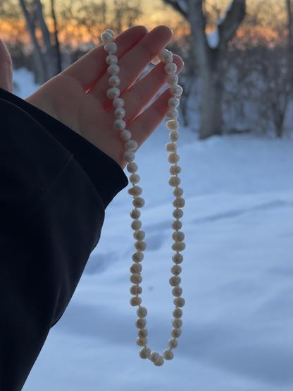 A hand in a black coat sleeve holding a knotted pearl necklace in front of a snowy backyard at sunset
