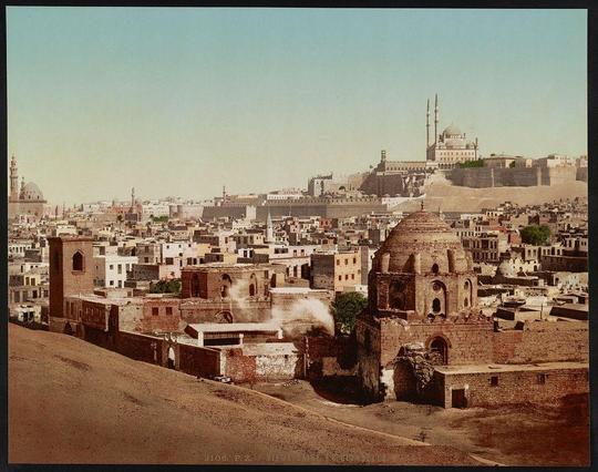 The image displays a panoramic view of the cityscape in Cairo, Egypt. It is an old photograph with a sepia tone that adds to its historical aura. The foreground shows a mix of buildings and structures varying from dilapidated remains on one side to well-preserved architectural elements like domes, archways, and minarets indicative of Islamic architecture.

In the midground lies Cairo's dense urban sprawl, densely packed with smaller houses or tenements that are typical for an old city. Among these residential buildings stand prominent historical structures such as mosques featuring distinctive Islamic features like pointed arches in windows, domes, and graceful minarets rising into a partly cloudy sky.

The background is dominated by the Citadel of Saladin (Citadel Hill), which overlooks much of Cairo's urban expanse. Visible are parts of its fortifications with walls that extend across what appears to be an arid landscape hinting at Egypt's desert proximity. The presence of smoke or steam from a chimney amidst these structures adds a dynamic element, suggesting life and activity within the city.

The overall composition suggests this photograph was taken for educational or documentary purposes, capturing not just Cairo but also offering insights into its historical layers as it stood around the turn of the 20th century.