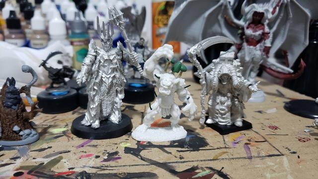 Mini painting desk, with paint stained cardboard below a bunch of minis. In front are a metal mini of Sauron on the left, a white plastic troll with wires sticking out of it, and a large orc in a heavy cloak with a backpack full of books, holding a scythe in one hand
