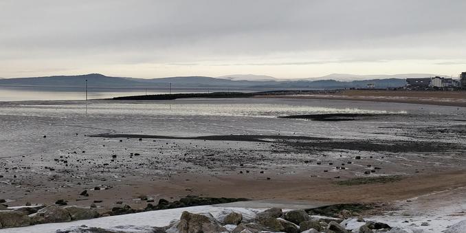 An almost monochrome picture of Morecambe Bay.

The hills of the Lake District are a dark line on the horizon, the snow-covered tops merging with the cloudy sky. It's high tide, the sea is a thin line of grey behind an expanse of iron-grey mud. A dark peninsula juts out into the mud. In the foreground are some dark grey boulders spattered with snow.