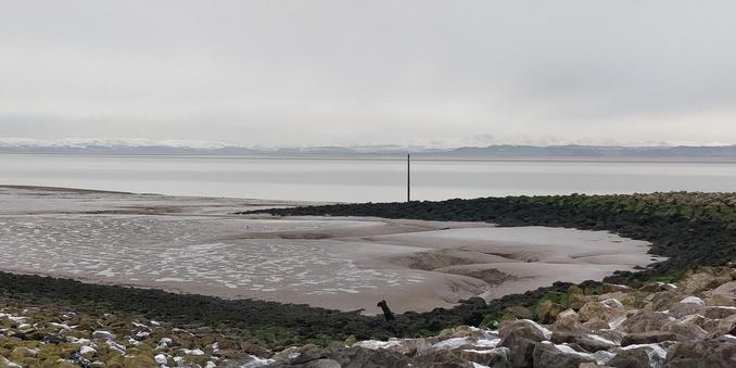 An almost monochrome picture of Morecambe Bay.

The hills of the Lake District are a pale line on the horizon, the snow-covered tops merging with the cloudy sky. It's high tide, the sea is a band of light grey behind an expanse of iron-grey mud. A stone groyne sweeps across the foreground and curves out into the mud, its dark grey boulders spattered with snow.