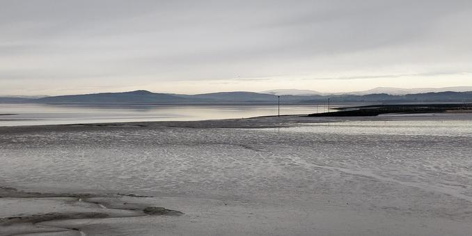 An almost monochrome picture of Morecambe Bay.

The hills of the Lake District are a dark line on the horizon, the snow-covered tops merging with the cloudy sky. It's high tide, the sea is a band of light grey behind an expanse of iron-grey mud. A stone groyne cuts its way horizontally across the mud.
