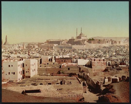 The image depicts a vast urban landscape, likely captured in the late 19th or early 20th century. Dominating the skyline is an expansive fortified complex with multiple minarets and domes, indicative of Islamic architecture. This could be identified as part of Cairo's historical citadel, known for its blend of ancient fortifications and significant religious buildings.

The cityscape includes a mix of dense urban structures, showcasing various architectural styles from different periods. Some buildings appear older, possibly centuries old with exposed brickwork and smaller domes, while others show more modern construction techniques.

The coloration suggests the image might be an original photograph or a print made on tinted paper, common in photographic prints during that era. The sky is clear blue above the city, hinting at good weather conditions when the photo was taken.

In terms of geographical and historical context, this landscape corresponds to Cairo's Mokattam district as referenced by "Vieux Caire" (Old Cairo), which holds significant archaeological sites like the ancient village of Babylon. The caption also mentions Zürich: Photoglob Company, indicating that this image may be part of a series created or distributed in Switzerland during the period mentioned.

Given these details, it's plausible to deduce that the photograph captures a perspective from an elevated position overlooking Cairo’s Mokattam district [...]