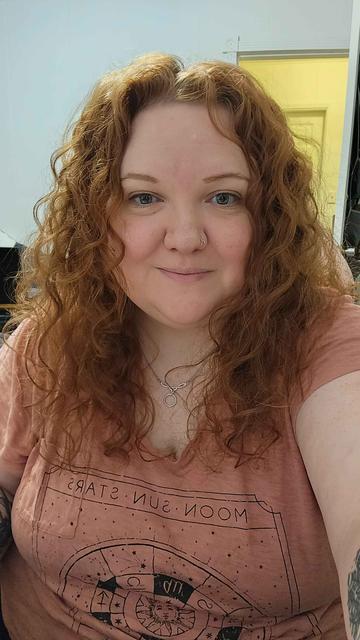 Me, a red haired white woman, smiling into the camera. My hair is curly with a bit of volume and some frizz.
