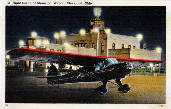 Nighttime falls over the airport. You can see the lights of the tower and airport in the background. A dark-colored plane is in the foreground with engine spinning about to taxi away