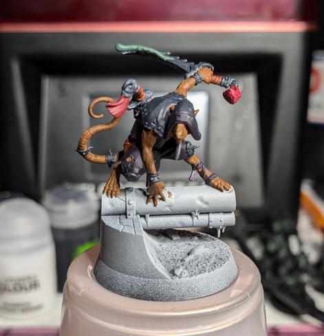 A miniature of a ninja rat hero posing on pipe in a sewer. It has a simple basecoat of tan skin, deep purple pants and a black hood. It's carrying two swords with red tassels on the ends and dripping green poison.