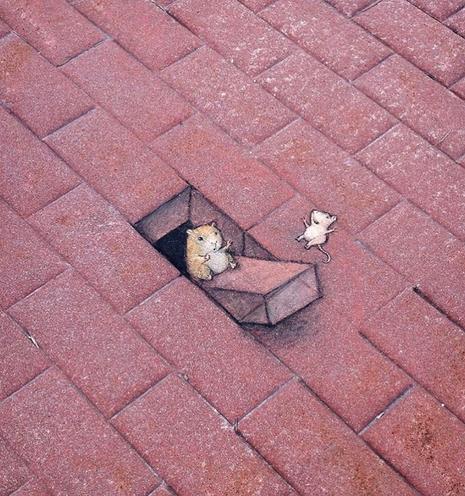 Streetart. A small mouse, a hamster and a single cobblestone have been painted with chalk and 3D effect on a sidewalk with red cobblestones. The "painted" cobblestone sticks out of a hole because a small, fat hamster is sitting at one end. He tilts the stone like a seesaw so that a little mouse (called a tit) at the other end can fly happily through the air and have fun.