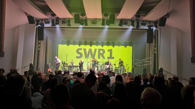 Das Bild zeigt eine Live-Konzertszene mit einer Band, die auf der Bühne spielt. Es sind sieben Personen auf der Bühne zu sehen, die Musikinstrumente oder Mikrofone halten. Im Hintergrund dominiert ein großer Bildschirm mit den Buchstaben "SWR1", die in Weiß auf einem grünen Hintergrund leuchten. Vor der Bühne steht ein Publikum, von dem einige Personen zu sehen sind, die den Darstellern zugewandt sind. Die Beleuchtung ist im Publikumsbereich gedimmt und auf die Bühne gerichtet, um die Darsteller hervorzuheben.