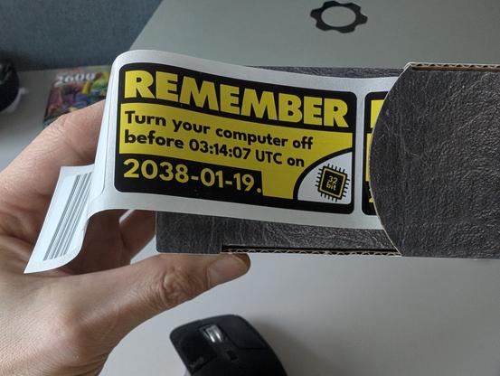 A hand holding a sticker dispensers.
One sticker is visible. It's yellow/black/white. In the lower right corner is a stylized 32bit microchip. The text reads:

REMEMBER
Turn your computer off before 03:14:07 UTC on 2038-01-19.