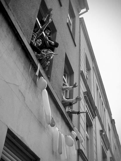 das schwarzweißfoto im hochformat zeigt feiernde und verkleidete menschen, die während eines karnevalsumzugs im und am fenster stehen und ihre arme ausstrecken.
vom bürgersteig nach oben in den 1. stock fotografiert sind zwei fenster eines mehrgeschossigen hauses zu sehen. in beiden fenstern stehen verkleidete junge menschen, die scheinbar auf die straße schauen, rufen und dabei ihre hände und arme ausstrecken.
unterhalb beider fenster ist eine schnur mit ballons befestigt.