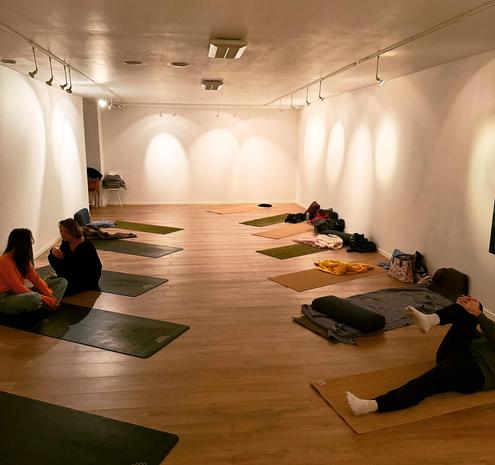 This image depicts an indoor yoga or meditation studio with wooden floors and white walls. The space is softly lit with overhead lights, creating a calm and cozy atmosphere. Yoga mats are spread across the floor in an organized manner, with some mats having blankets, bolsters, and personal items like bags and jackets placed next to them.

A few individuals are visible in the room. Two people seated on one of the mats near the left side appear to be engaged in conversation. Another person lies on their back on a mat on the right side, stretching one leg. The room feels peaceful and welcoming, likely in preparation for a yoga or relaxation session.