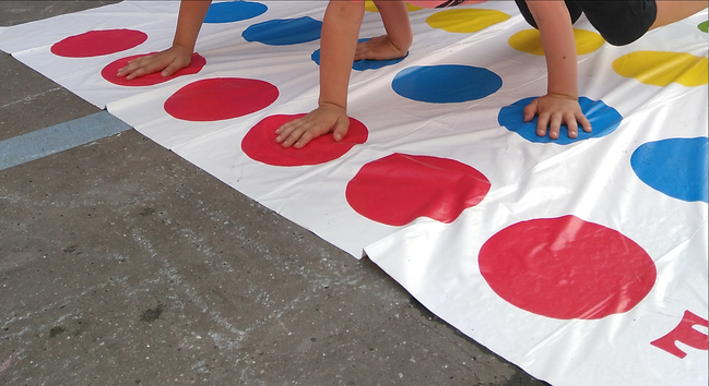 Dans une cour bitumée, tapis de Twister avec des cercles colorés, sur lesquels sont posées des mains d'enfant
