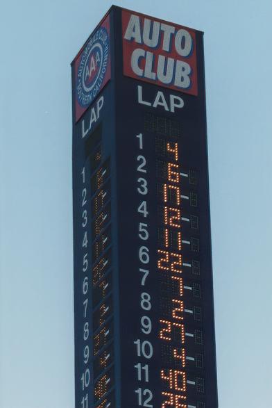 A tall digital scoring pylon displaying race positions and driver numbers, branded with "AUTO CLUB" and "LAP." The background is a blue sky.