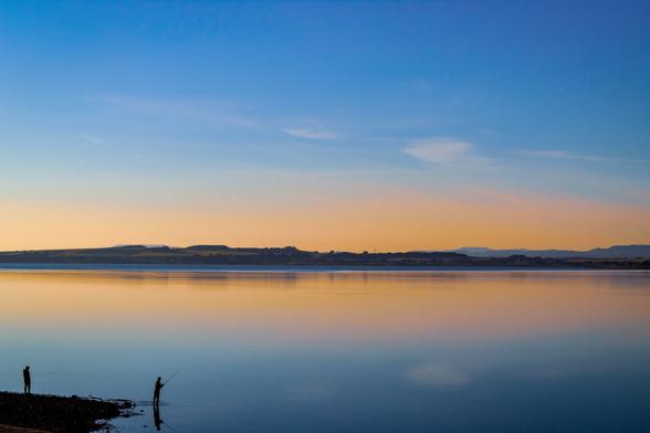 Landscape of a big lake with some small hills on the horizon and in the foreground two silhouettes, one of them is fishing.