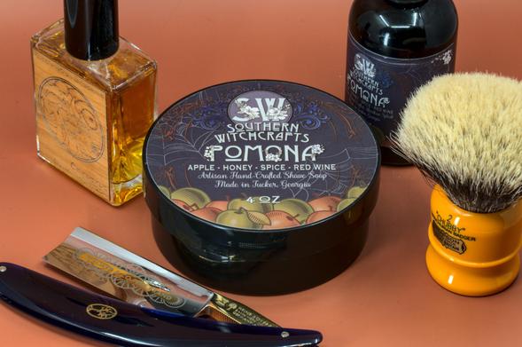 (ChatGPT description)

This image showcases an artisanal wet shaving setup featuring products from Southern Witchcrafts' Pomona line, which is inspired by warm, autumnal scents. The setup exudes a sophisticated and seasonal aesthetic, perfect for enthusiasts of luxurious and themed grooming rituals.

At the center is a 4 oz tub of Pomona shaving soap, with a beautifully detailed label depicting apples, vines, and floral elements, highlighting the scent profile of apple, honey, spice, and red wine. This artisan-crafted soap, made in Tucker, Georgia, promises a rich, indulgent lather and a warm, cozy fragrance.

To the right of the soap, a matching bottle of Pomona aftershave splash continues the theme with its deep amber tones and complementary label design. The aftershave provides a soothing finish to the shave, enhancing the lingering scent of Pomona’s unique blend.

On the left, a striking bottle of cologne or aftershave splash with a wooden label adds a touch of craftsmanship and earthiness to the setup. The intricate engraving hints at the product's artisanal quality, making it a perfect companion to the Pomona line.

In the foreground, a straight razor with a dark blue handle and gold-etched blade completes the setup. Its polished, sleek design emphasizes precision and tradition, adding a timeless elegance to the arrangement.

To the far right is the renowned Chubby 2 shaving brush, with its bright orange handle and dense badger bristles.