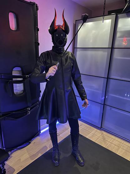 Loki in a long leather dress and boots, wearing a full face black gas mask and a set of light up Mercy Imp horns from Overwatch. They’re holding a whip menacingly.