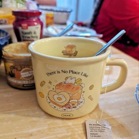 Frühstückstisch, im Fokus eine hellgelbe Tasse mit einem gemalten Häschen hinter einem Toastbrot und eine Tasse Tee, umgeben von weiterem Gebäck. Daneben liegt eine herzförmige Brotscheibe auf dem Bild mit einem Spiegelei. Das Bild ist umrahmt mit den Worten "There is no place like home"