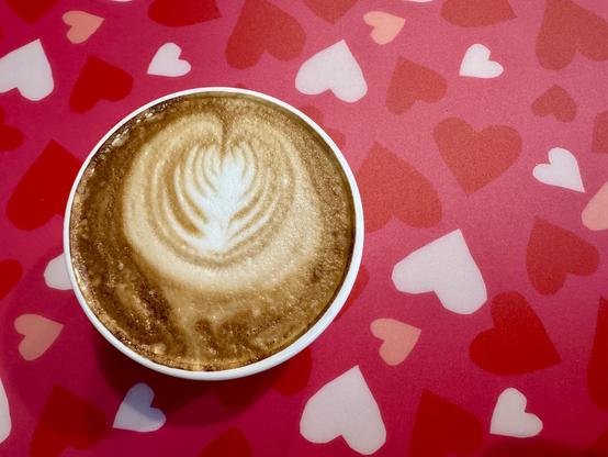 Overhead picture of a small white mug filled with a cappuccino sitting on pink background with lots of different shaded hearts.