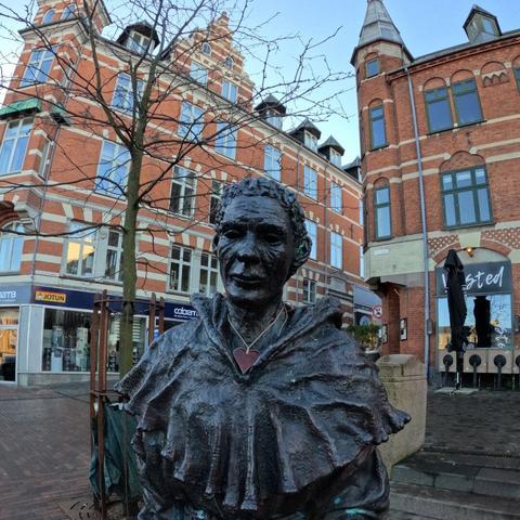 Top half of a statue in dark metal of a monk with a red heart necklace in ceramic around the neck. The statue stands in an open space in front of a couple of posh city houses in red brick, with a single tiny tree to the left. I would say the monk looks rather pleased.