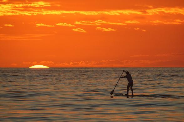 A tip of a yellow rising sun  can be seen on the horizon, on the seafront. There is a stand-up-paddler in the foreground. Colors are mostly orange tones.