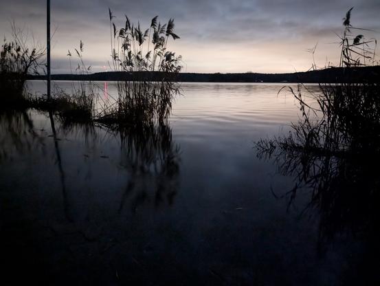 Written with the help of Claude:

Atmospheric twilight shot on the Havel in the 'blue hour'. The special feature: The flooded riverside path has completely merged with the river, blurring the boundaries between land and water. In the foreground, dark reed stalks protrude from the mirror-smooth water surface, their silhouettes casting long shadows. The surface of the water stretches seamlessly to the horizon, where a chain of wooded hills emerges as a dark line. In the pastel-coloured evening sky, the sun breaks through the cloud cover as a dull, red dot and draws a narrow, reddish reflection on the water. The muted blue and grey tones of the water merge with the sky, reinforcing the boundlessness of the flood. The entire scene conveys an almost surreal atmosphere in which the familiar structures of the landscape dissolve into a large, calm expanse of water.